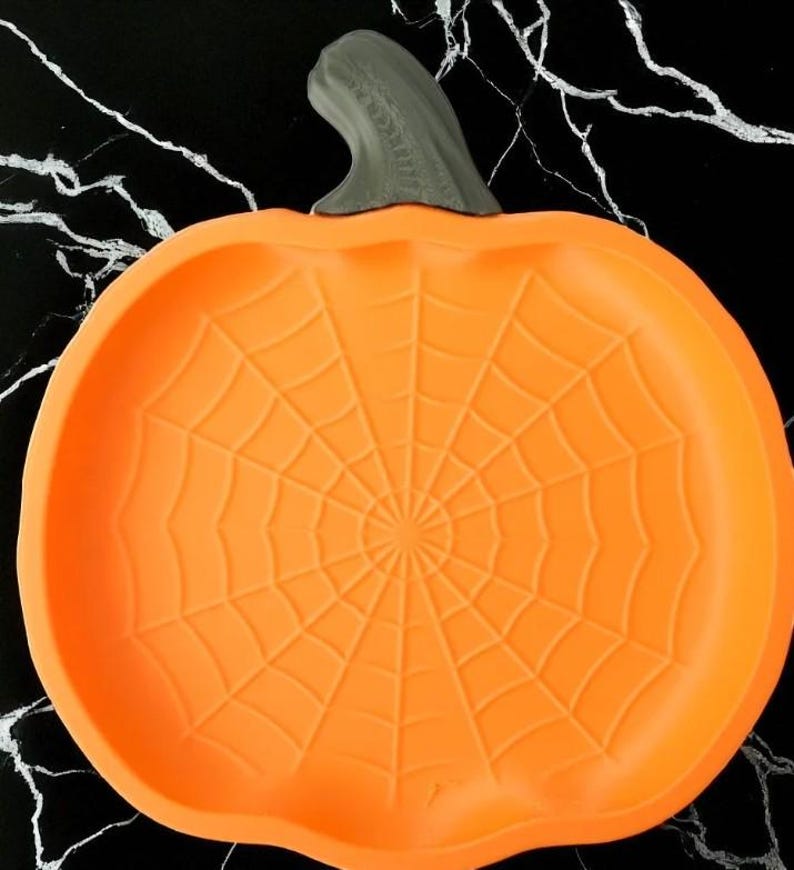 May include: Orange pumpkin-shaped serving dish with a gray stem. The dish features a spiderweb design in the center, perfect for Halloween. The dish is made of a flexible material.