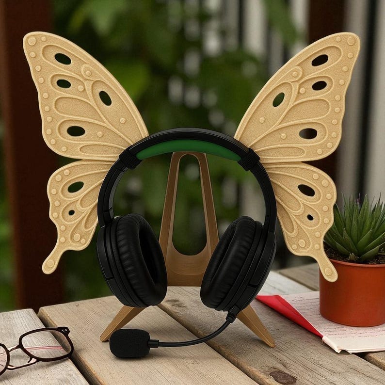 May include: Black headphones with a green headband and a microphone sit on a wooden butterfly wing-shaped stand. The wings are beige with decorative cutouts. A small potted plant and eyeglasses are in the foreground.