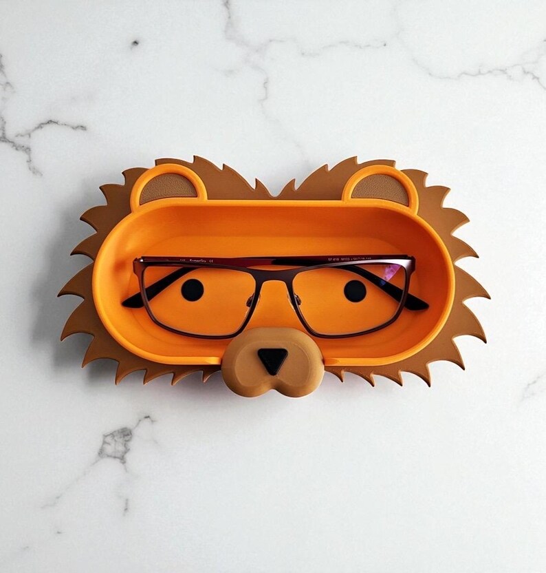 May include: A novelty glasses holder in the shape of a lion, featuring an orange tray for glasses and a brown mane. The holder contains a pair of black-framed glasses. The design includes a lion's nose.