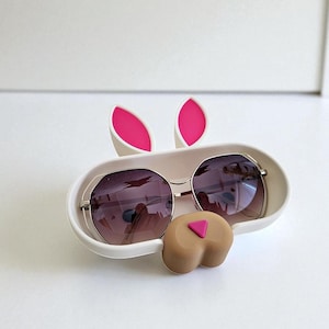 May include: A white bunny-shaped sunglasses holder with pink accents. The holder features a pair of sunglasses with a silver frame and dark lenses. The bunny design includes pink ears and a brown nose with a pink triangle.
