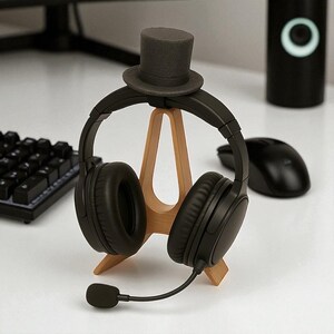 May include: Black over-ear headphones with a boom microphone rest on a light brown wooden stand. A small, gray top hat sits atop the headphones. A black computer mouse and keyboard are visible in the background.
