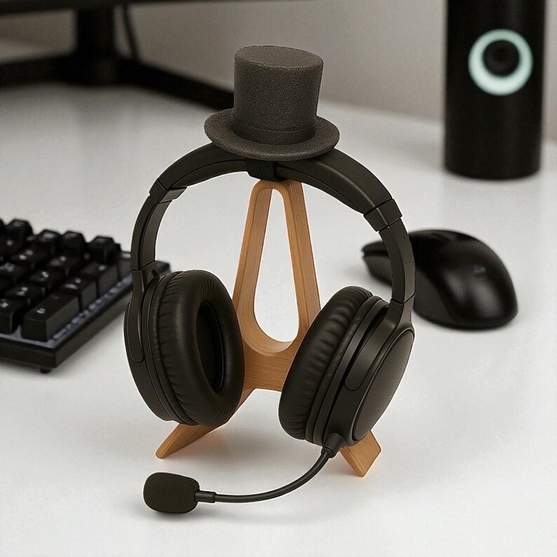 May include: Black over-ear headphones with a boom microphone rest on a light brown wooden stand. A small, gray top hat sits atop the headphones. A black computer mouse and keyboard are visible in the background.