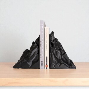 May include: Black mountain-shaped bookends holding two books. The bookends have a textured surface and are made of a solid material. The books are titled "The Strength in Our Scars" and "&Uuml;berwintern".
