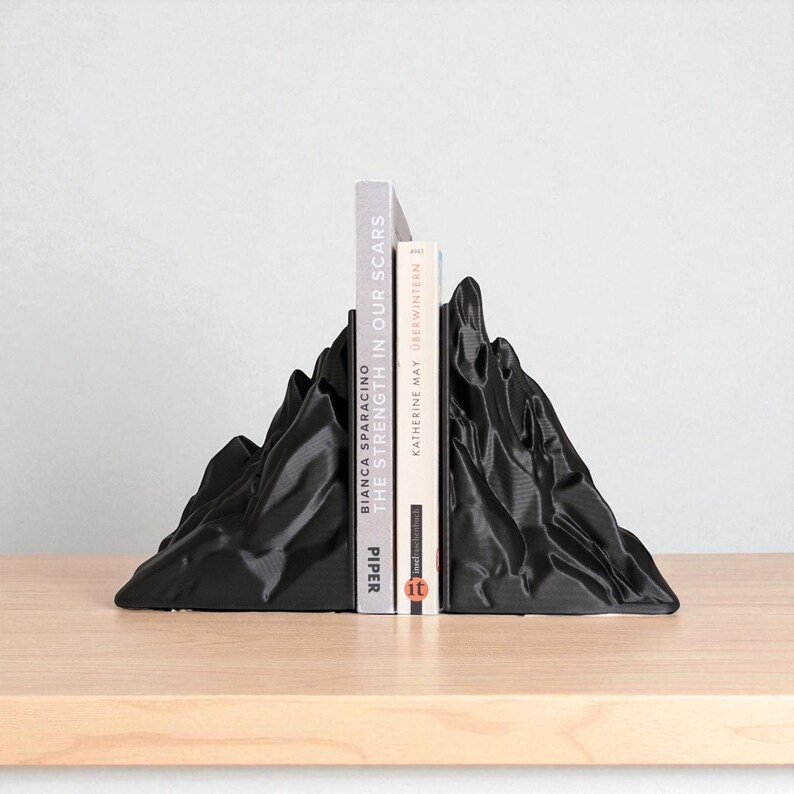May include: Black mountain-shaped bookends holding two books. The bookends have a textured surface and are made of a solid material. The books are titled "The Strength in Our Scars" and "&Uuml;berwintern".