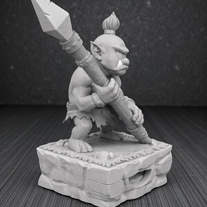 May include: A gray figurine of a fantasy creature holding a spear. The figure is standing on a stone base with a textured surface. The creature has a large nose, pointed ears, and a tuft of hair on its head.