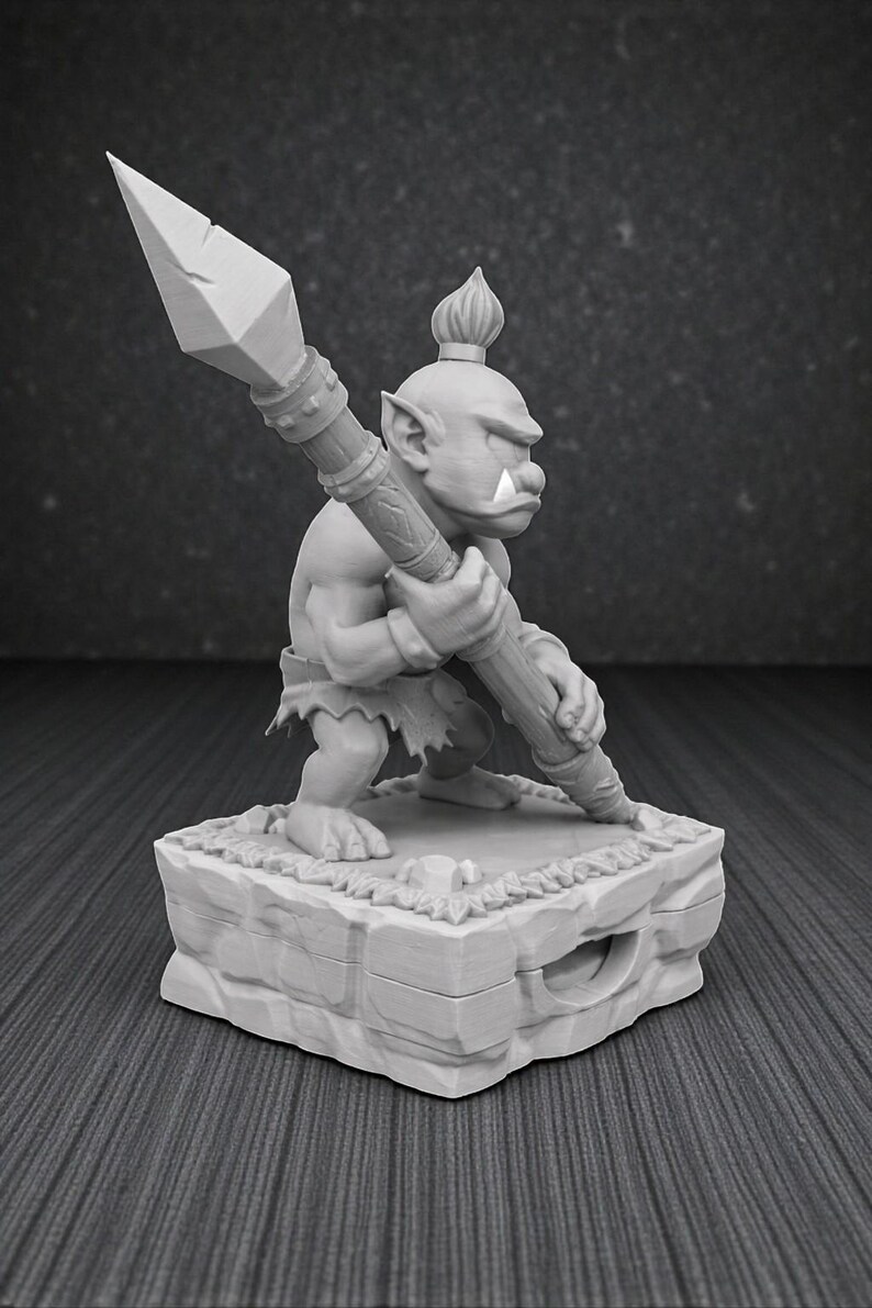 May include: A gray figurine of a fantasy creature holding a spear. The figure is standing on a stone base with a textured surface. The creature has a large nose, pointed ears, and a tuft of hair on its head.