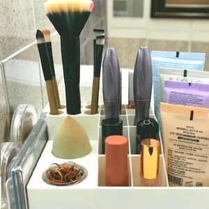 May include: A white makeup organizer filled with various beauty products. Includes makeup brushes, mascara tubes, a makeup sponge, and tubes of skincare products. The brushes have black handles and colorful bristles. The organizer is on a bathroom counter.
