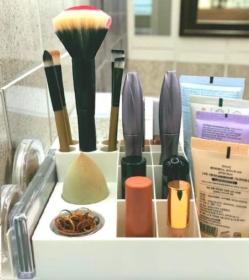 May include: A white makeup organizer filled with various beauty products. Includes makeup brushes, mascara tubes, a makeup sponge, and tubes of skincare products. The brushes have black handles and colorful bristles. The organizer is on a bathroom counter.