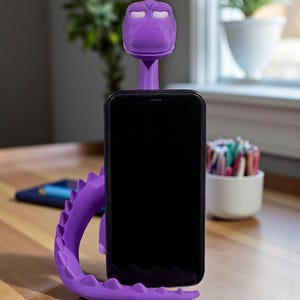 May include: A purple dragon-shaped phone stand holds a black smartphone. The stand is made of a 3D-printed material and has a detailed design. The dragon's head supports the phone, and its tail curves around the back. The stand is on a wooden surface.