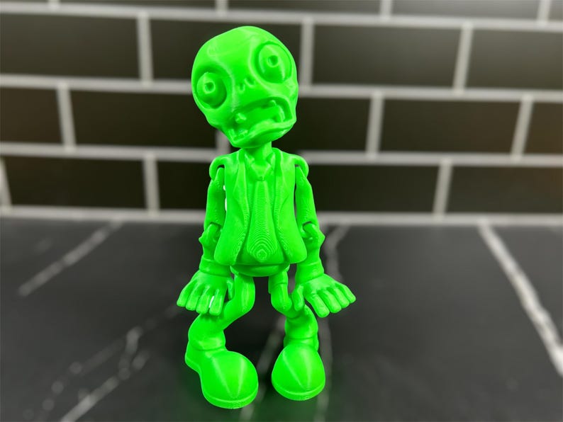 May include: A bright green 3D printed figurine of a zombie wearing a suit and tie. The zombie has a wide open mouth and large eyes.