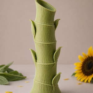 Puede incluir: Un jarr&oacute;n verde con forma de bamb&uacute; con una superficie texturizada y detalles de hojas. El jarr&oacute;n tiene una abertura inclinada y est&aacute; rodeado de un girasol y p&eacute;talos amarillos dispersos. El jarr&oacute;n mide unos 25 cm de alto.