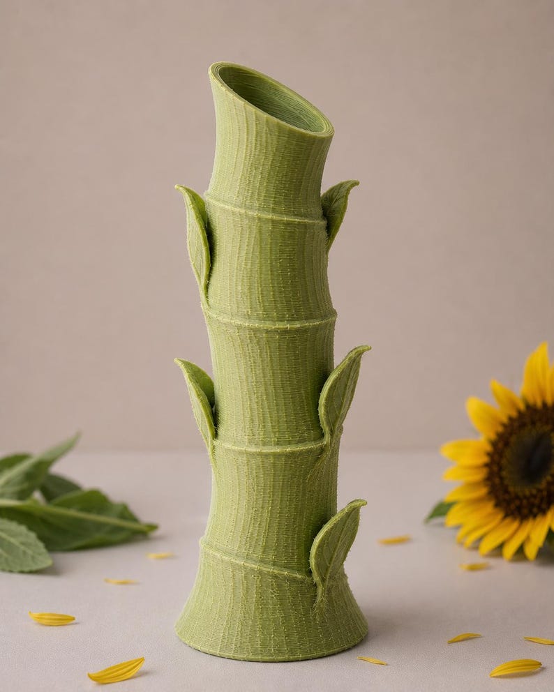 Puede incluir: Un jarr&oacute;n verde con forma de bamb&uacute; con una superficie texturizada y detalles de hojas. El jarr&oacute;n tiene una abertura inclinada y est&aacute; rodeado de un girasol y p&eacute;talos amarillos dispersos. El jarr&oacute;n mide unos 25 cm de alto.
