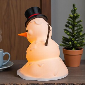 May include: A snowman-shaped lamp with a black top hat and an orange carrot nose. The lamp emits a warm, glowing light and has stick arms and black button eyes. A small potted evergreen tree is next to it.