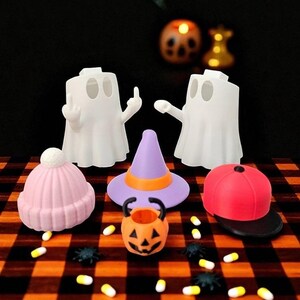 May include: A Halloween-themed display featuring white ghost figures, a pink beanie, a purple witch hat with an orange band, and a red baseball cap. An orange jack-o'-lantern bucket and scattered candies and spiders complete the festive scene.
