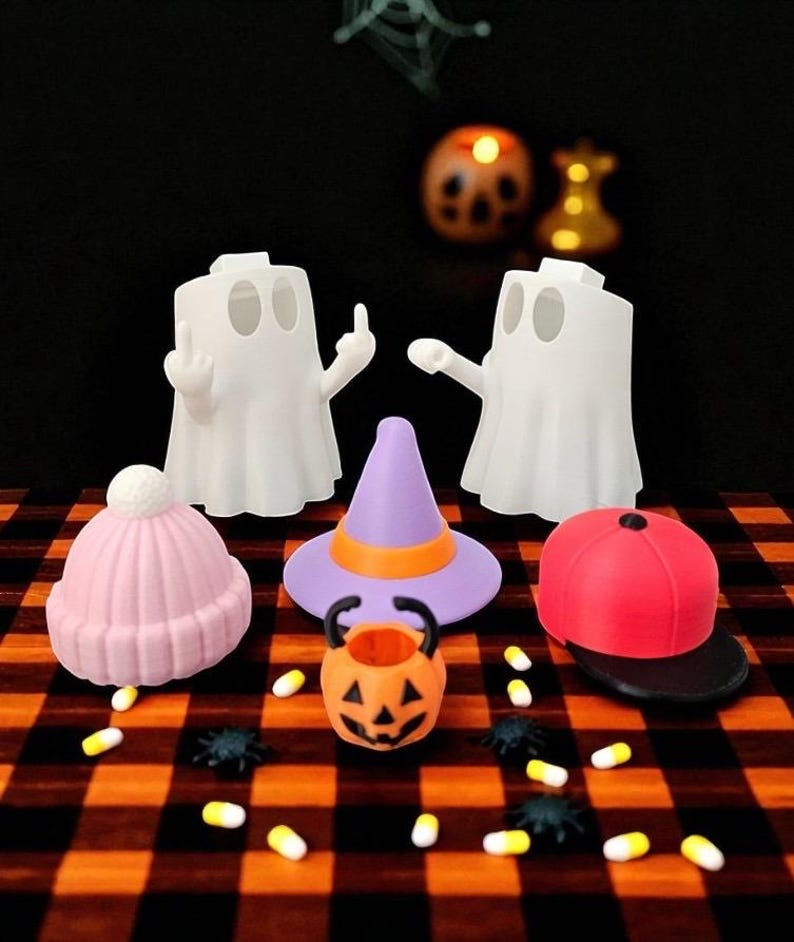 May include: A Halloween-themed display featuring white ghost figures, a pink beanie, a purple witch hat with an orange band, and a red baseball cap. An orange jack-o'-lantern bucket and scattered candies and spiders complete the festive scene.