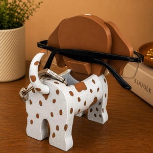 May include: A white and brown spotted dog-shaped desk organizer. The organizer holds eyeglasses and keys. The dog has brown ears and spots. The eyeglasses are black-framed. The organizer is made of a speckled material.