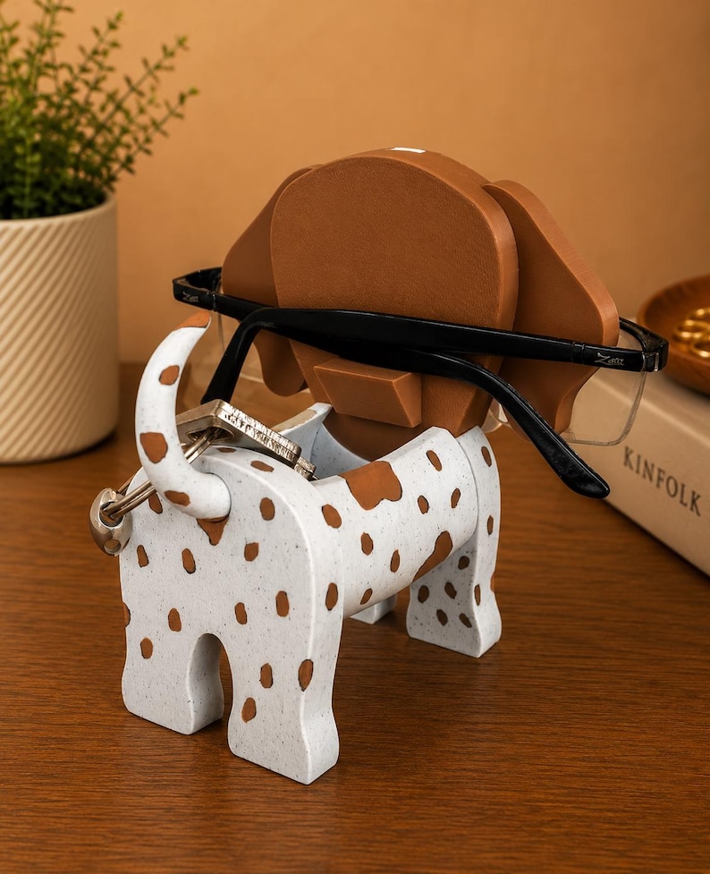 May include: A white and brown spotted dog-shaped desk organizer. The organizer holds eyeglasses and keys. The dog has brown ears and spots. The eyeglasses are black-framed. The organizer is made of a speckled material.