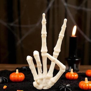 May include: A cream-colored skeleton hand sculpture in a peace sign gesture. The hand is surrounded by miniature orange pumpkins, black spiders, and a lit black candle, creating a Halloween theme. The background is dark.