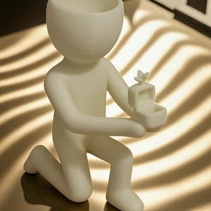 May include: A 3D-printed figurine of a person kneeling, holding a ring box. The figure is white and has a smooth texture. The ring box is open, with a small butterfly on top. The figurine is a decorative item.