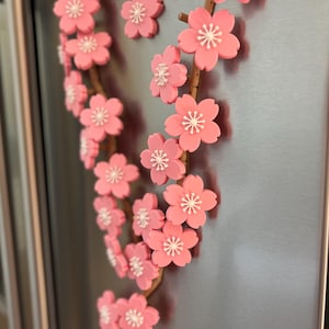 May include: A decorative floral branch with pink cherry blossoms and white centers. The branch is brown and appears to be made of a flexible material. The blossoms are attached to a metallic surface, likely a refrigerator or appliance.