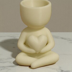 May include: A cream-colored figurine of a person in a seated yoga pose, holding a heart-shaped object. The figurine has a hollowed-out head, designed to hold small plants or items. The figurine is on a white and gray marble surface.