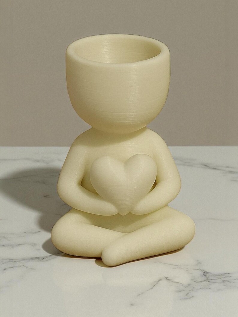 May include: A cream-colored figurine of a person in a seated yoga pose, holding a heart-shaped object. The figurine has a hollowed-out head, designed to hold small plants or items. The figurine is on a white and gray marble surface.