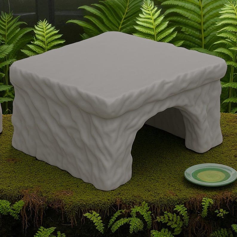 May include: A light gray, stone-like pet habitat with a rectangular top and an arched opening. The textured surface resembles carved stone. A small, round, green dish sits nearby. The habitat is on a bed of green moss.