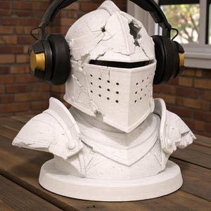 Puede incluir: Busto de casco de caballero blanco con auriculares negros y dorados. El casco tiene un aspecto desgastado y agrietado. Los auriculares son negros con detalles dorados en los auriculares. El busto est&aacute; sobre una base redonda.