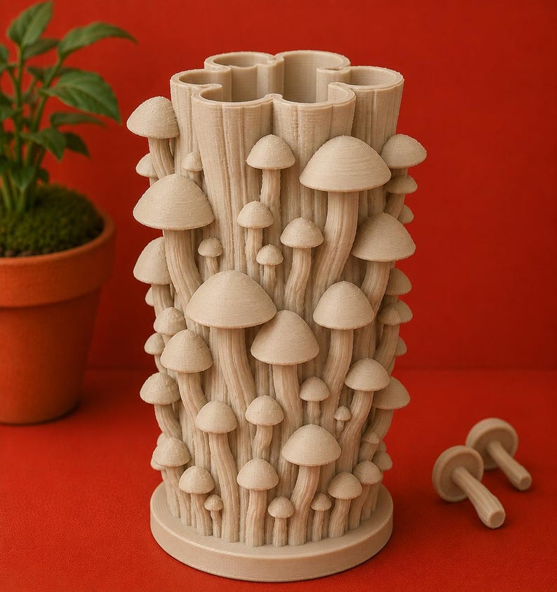 K&ouml;nnte beinhalten: Eine beige Vase mit Pilz-Motiv und strukturierter Oberfl&auml;che. Die Vase ist mit Pilzkappen und -stielen in verschiedenen Gr&ouml;&szlig;en bedeckt. Zwei zus&auml;tzliche pilzf&ouml;rmige Objekte befinden sich neben der Vase. Die Vase steht auf einer roten Oberfl&auml;che.