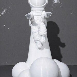 Op de afbeelding: Een witte astronautenfiguur in een zwevende pose, met uitgestrekte armen. De astronaut staat op een witte wolkachtige basis. De achtergrond is effen grijs met witte spikkels, wat een ruimtelijke esthetiek cre&euml;ert.