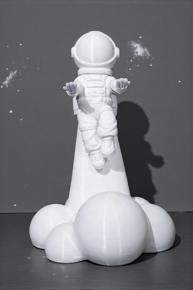 Op de afbeelding: Een witte astronautenfiguur in een zwevende pose, met uitgestrekte armen. De astronaut staat op een witte wolkachtige basis. De achtergrond is effen grijs met witte spikkels, wat een ruimtelijke esthetiek cre&euml;ert.