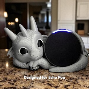 May include: A gray dragon-shaped speaker holder cradling a black Echo Pop speaker. The dragon has large eyes and a friendly expression. The text "Designed for Echo Pop" is printed at the bottom.