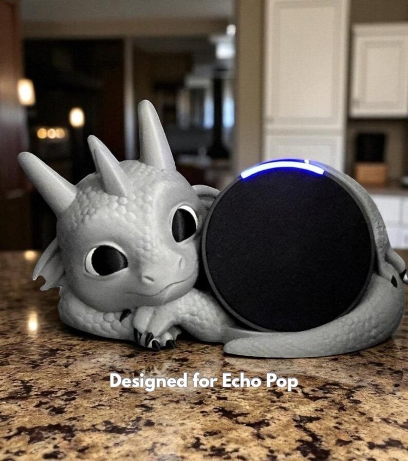 May include: A gray dragon-shaped speaker holder cradling a black Echo Pop speaker. The dragon has large eyes and a friendly expression. The text "Designed for Echo Pop" is printed at the bottom.