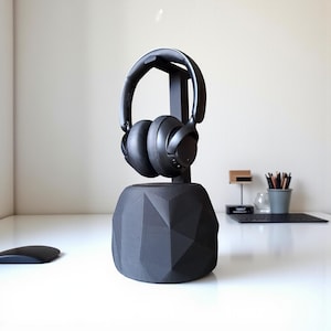 May include: Black over-ear headphones on a black geometric headphone stand. The stand has a faceted design and a vertical support. The headphones and stand are on a white surface. The stand is approximately 15 cm tall.