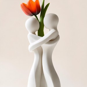 May include: A white ceramic vase featuring two embracing figures, holding a vibrant orange tulip with green leaves. The vase has a smooth, minimalist design, perfect for home decor. The tulip adds a pop of color against the white vase.
