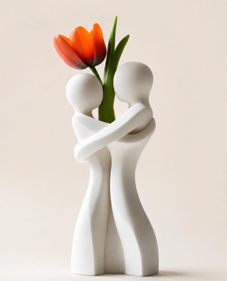 May include: A white ceramic vase featuring two embracing figures, holding a vibrant orange tulip with green leaves. The vase has a smooth, minimalist design, perfect for home decor. The tulip adds a pop of color against the white vase.