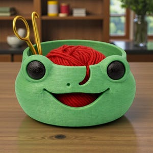 Könnte beinhalten: Eine grüne, froschförmige Garnschale mit einem roten Wollknäuel darin. Goldene Schere und Häkelnadel sind an der Seite platziert. Die Schale steht auf einer Holzoberfläche.