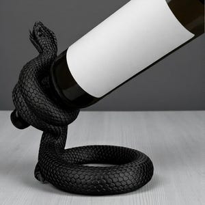 May include: A black snake wine bottle holder. The snake is coiled around the bottle, with its head raised. The bottle has a white label. The holder is on a light gray surface.