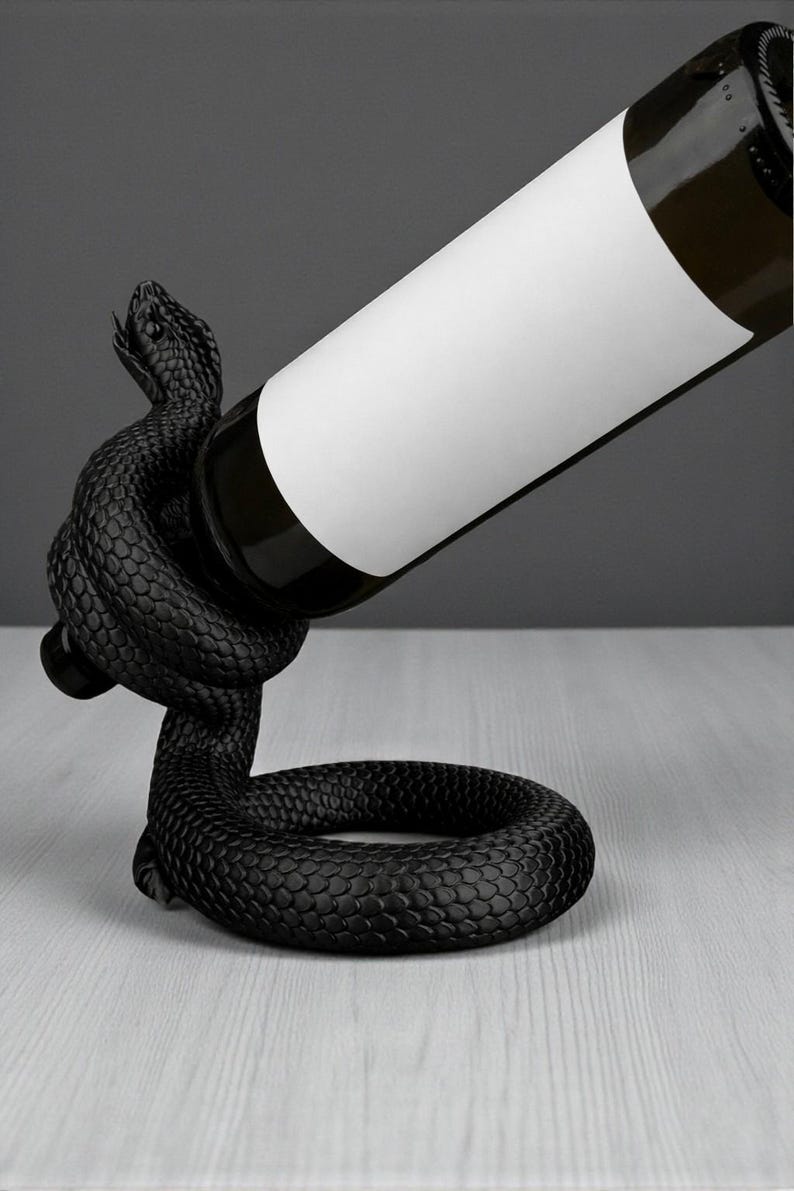 May include: A black snake wine bottle holder. The snake is coiled around the bottle, with its head raised. The bottle has a white label. The holder is on a light gray surface.