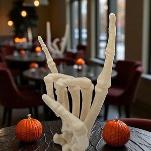 May include: A white, bone-colored sculpture of a hand making the rock-and-roll hand gesture. Two small orange pumpkins sit on either side of the sculpture. The background is a restaurant setting with tables and chairs.