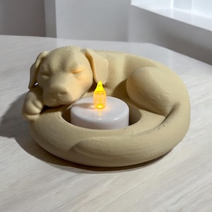 Puede incluir: Un portavelas beige con forma de perro con un dise&ntilde;o de perro durmiendo. El perro est&aacute; acurrucado alrededor de una vela blanca con una llama amarilla. El portavelas est&aacute; hecho de un material de color claro.
