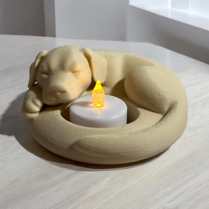 Puede incluir: Un portavelas beige con forma de perro con un dise&ntilde;o de perro durmiendo. El perro est&aacute; acurrucado alrededor de una vela blanca con una llama amarilla. El portavelas est&aacute; hecho de un material de color claro.