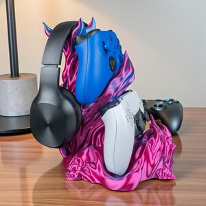 May include: A magenta and blue gaming controller and headset stand. The stand holds a black headset, a blue controller, and a white controller. The stand has a unique, flame-like design.
