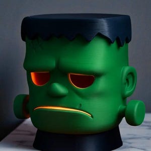 May include: A green Frankenstein-themed lamp with a black base and hair. The lamp has glowing orange eyes and mouth, and a stitched scar detail. The design is a stylized, cartoonish representation of the classic monster.
