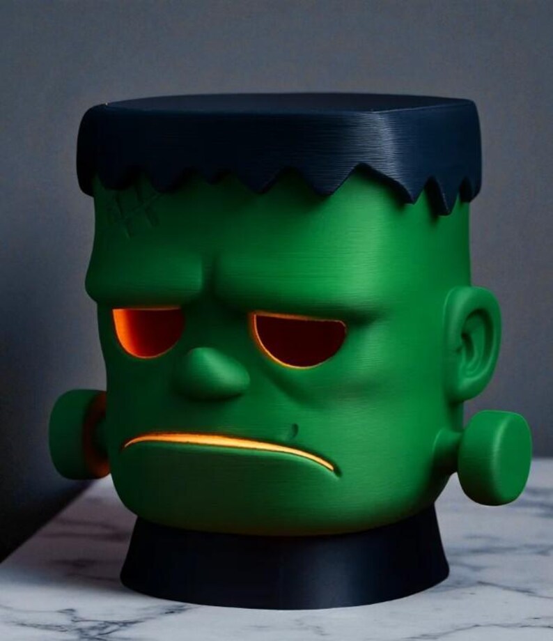 May include: A green Frankenstein-themed lamp with a black base and hair. The lamp has glowing orange eyes and mouth, and a stitched scar detail. The design is a stylized, cartoonish representation of the classic monster.