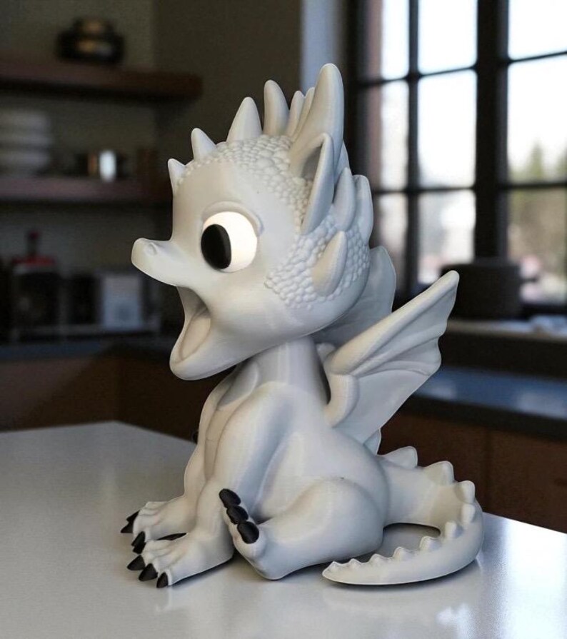May include: A white dragon figurine with large, expressive eyes and an open mouth. The dragon has detailed scales, wings, and black claws. It is sitting on a white surface, with a blurred background.