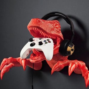 Könnte beinhalten: Roter, dinosaurierförmiger Wandhalter, der einen weißen Gamecontroller hält. Der Dinosaurier hat scharfe Zähne und Krallen, mit einem schwarzen Headset auf dem Kopf. Der Controller hat schwarze Tasten und farbige Richtungstasten.