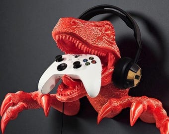 May include: A red dinosaur-shaped wall mount holding a white game controller. The dinosaur has sharp teeth and claws, with a black headset resting on its head. The controller has black buttons and colored directional buttons.
