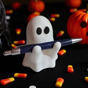May include: A white ghost-shaped pen holder with black eyes, holding a blue pen with silver accents. The Halloween-themed desk accessory is surrounded by candy corn and small pumpkins on a black surface.