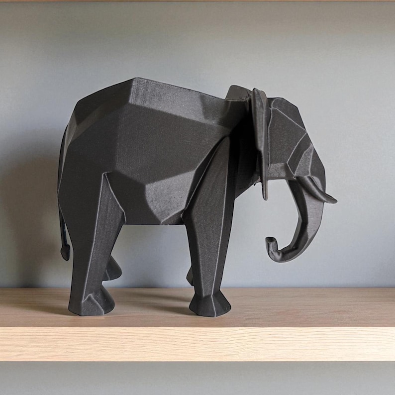Op de afbeelding: Een zwarte geometrische olifant sculptuur met een facetontwerp. De olifant heeft een slurf, slagtanden en grote oren. De sculptuur wordt tentoongesteld op een houten plank tegen een grijze muur. De olifant is een decoratief item.